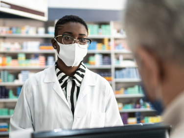 A pharmacist wearing a protective mask and white jacket talking to a customer.
