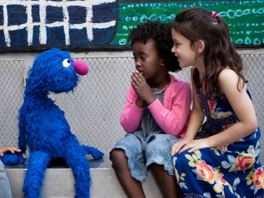 Two little girls talking to Sesame Street’s Grover on school bleachers.