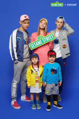 Three young adults pose behind two young children against a vibrant blue background. Everyone is wearing Sesame Street branded clothing.