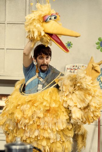 A young Caroll Spinney wears Big Bird's lower body. He holds Big Bird's head over his own head.