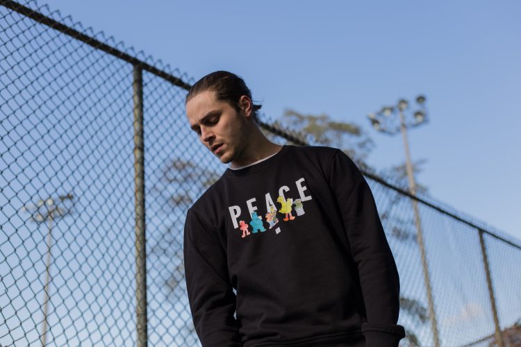 A model poses against a chain linked fence. He wears a long-sleeved black shirt with the word 