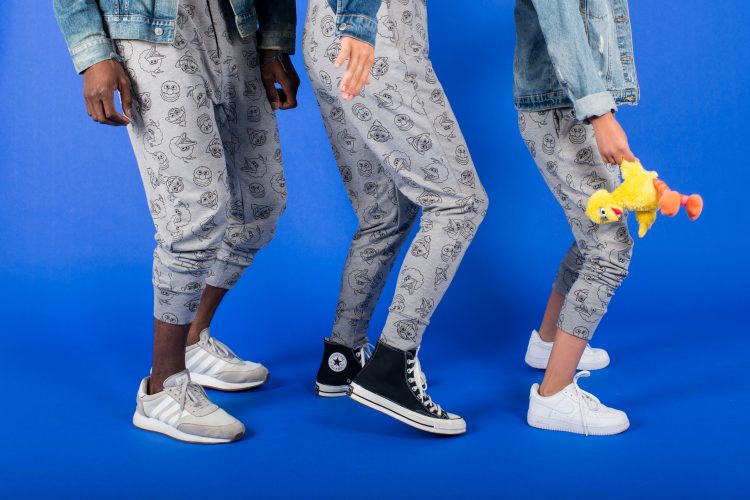A waist-down view of models showing off Sesame-branded pants. The pants are gray, athleisure-style, printed with line drawings of different Muppet heads.