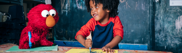 A young child smiles while coloring with Elmo.