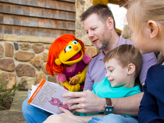 A father figure reads a book to his children as Muppet Julia looks on and smiles.