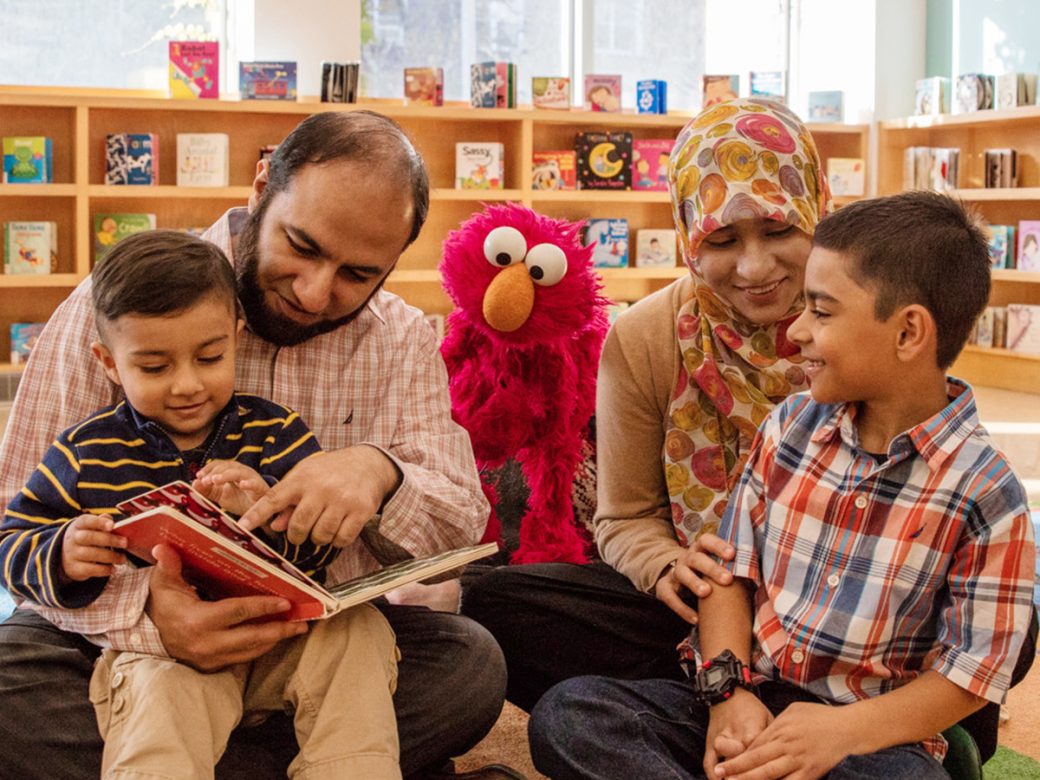 Parents reading to Elmo and their two kids in a library.