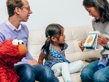 A young girl sits with Elmo and her family and reads flashcards.