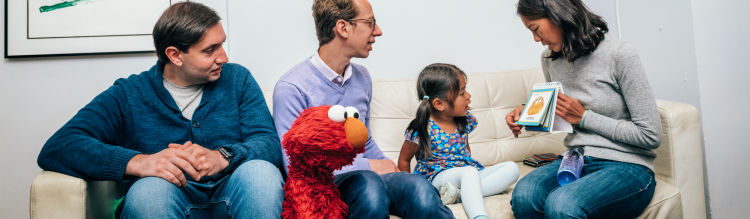 A young girl sits with Elmo and her family and reads flashcards.