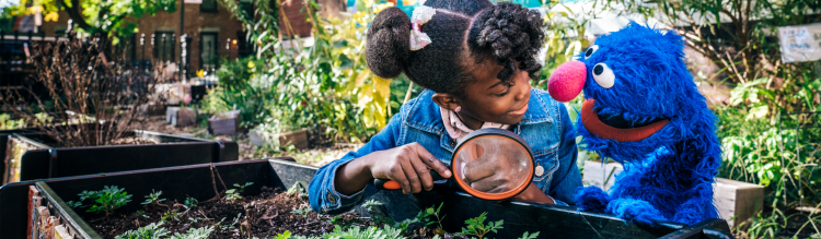 A young girl uses a magnifying glass to inspect soil with Grover.