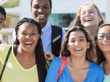 Eight young adults, some wearing backpacks, link arms and smile at us.