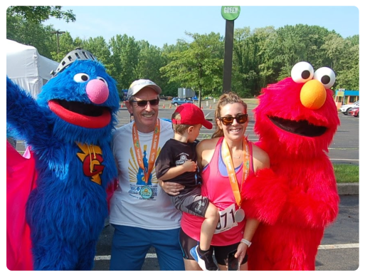 Two grown ups and a child pose in running gear and winner's medals between Grover and Elmo.