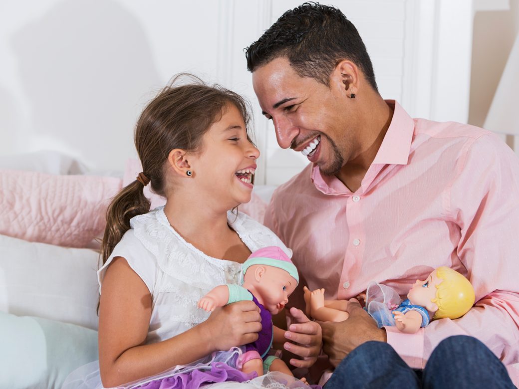 Father and young daughter laugh together, playing with dolls.