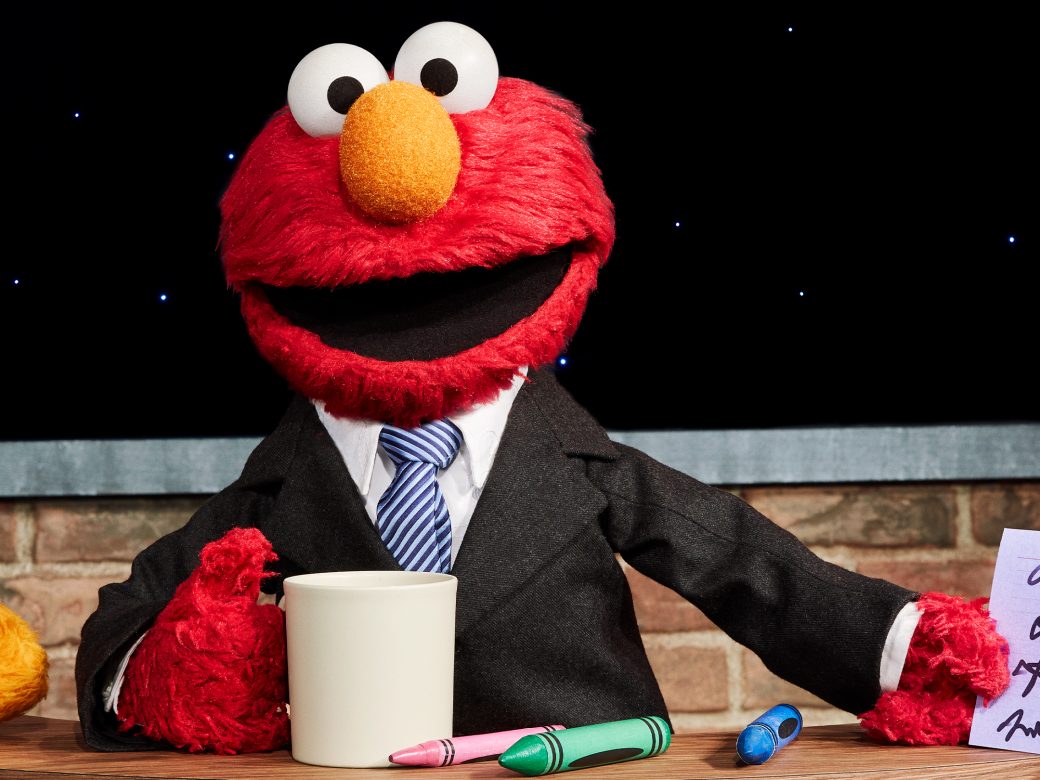 Elmo poses with a mug behind a desk wearing a suit.