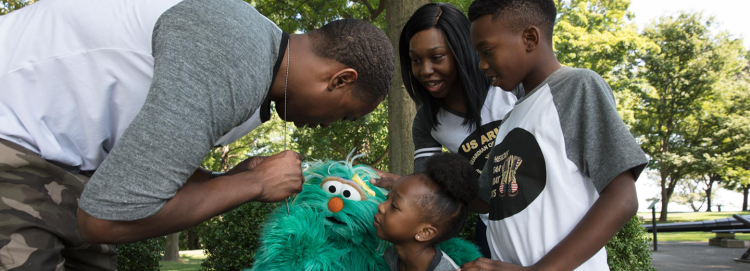 A father bends down to show his dogtags to Sesame Street’s Rosita and his family.