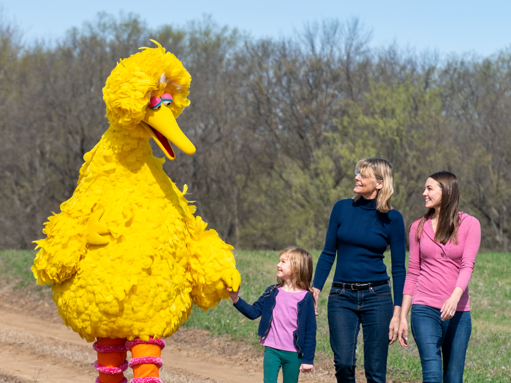 Big Bird walks on a dirt road with a young girl and two women.