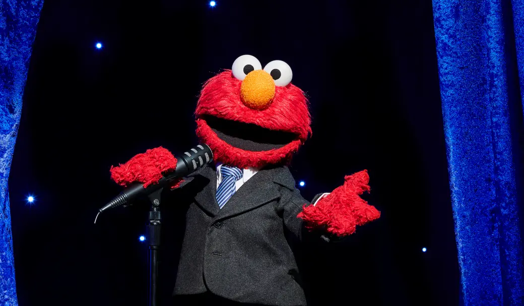 Elmo is dressed in a suit, holding a microphone and performing on stage.