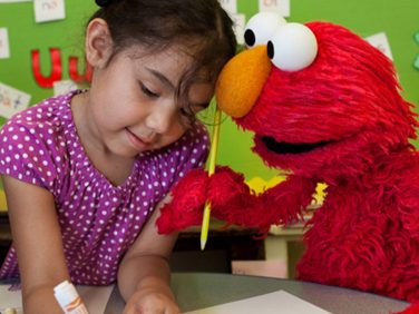Elmo with a pencil and a little girl at a table.