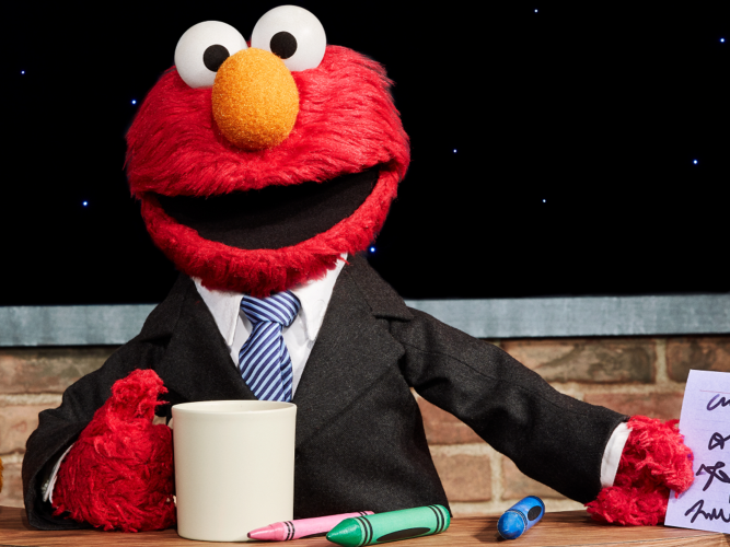 Elmo poses with a mug behind a desk wearing a suit.