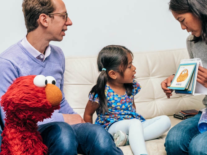Elmo sitting with a family on a couch as a parent reads aloud.