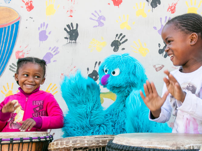 A smiling group of children play the drums alongside two happy Muppets from Takalani Sesame
