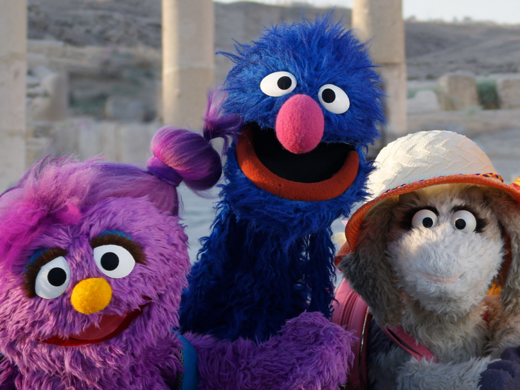 Group photo of four Muppets from Ahlan Simsim smiling and posing together amongst historic ruins.