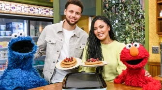 Stephen and Ayesha Curry each hold a plate of pancakes between Cookie Monster and Elmo.