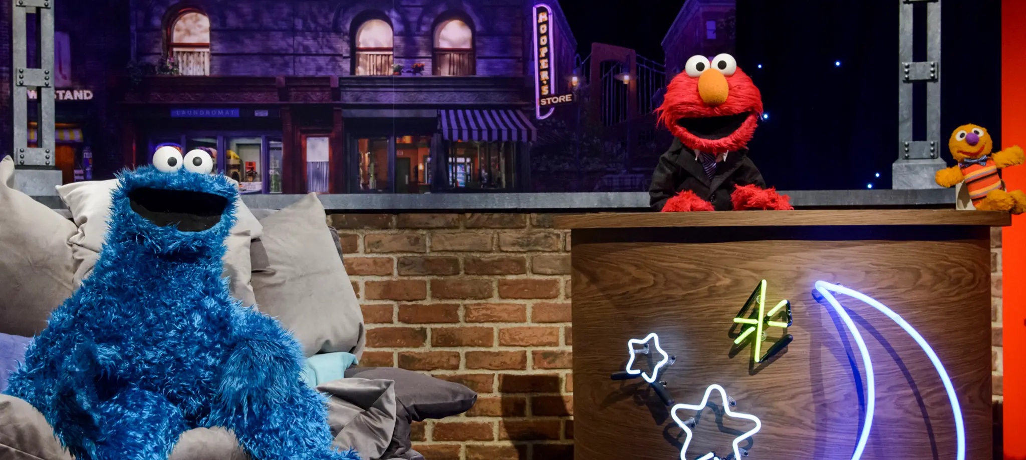 Elmo sits and a desk, and Cookie Monster sits next to him on a couch.