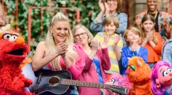 Kelsea Ballerini holds an acoustic guitar and claps. She's surrounded by muppets and neighbors.