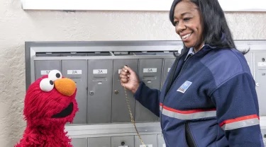 Elmo poses with a mail carrier smiling and opening a mailbox