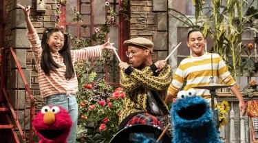 Elmo and Cookie Monster pose in front of an adult playing the drums