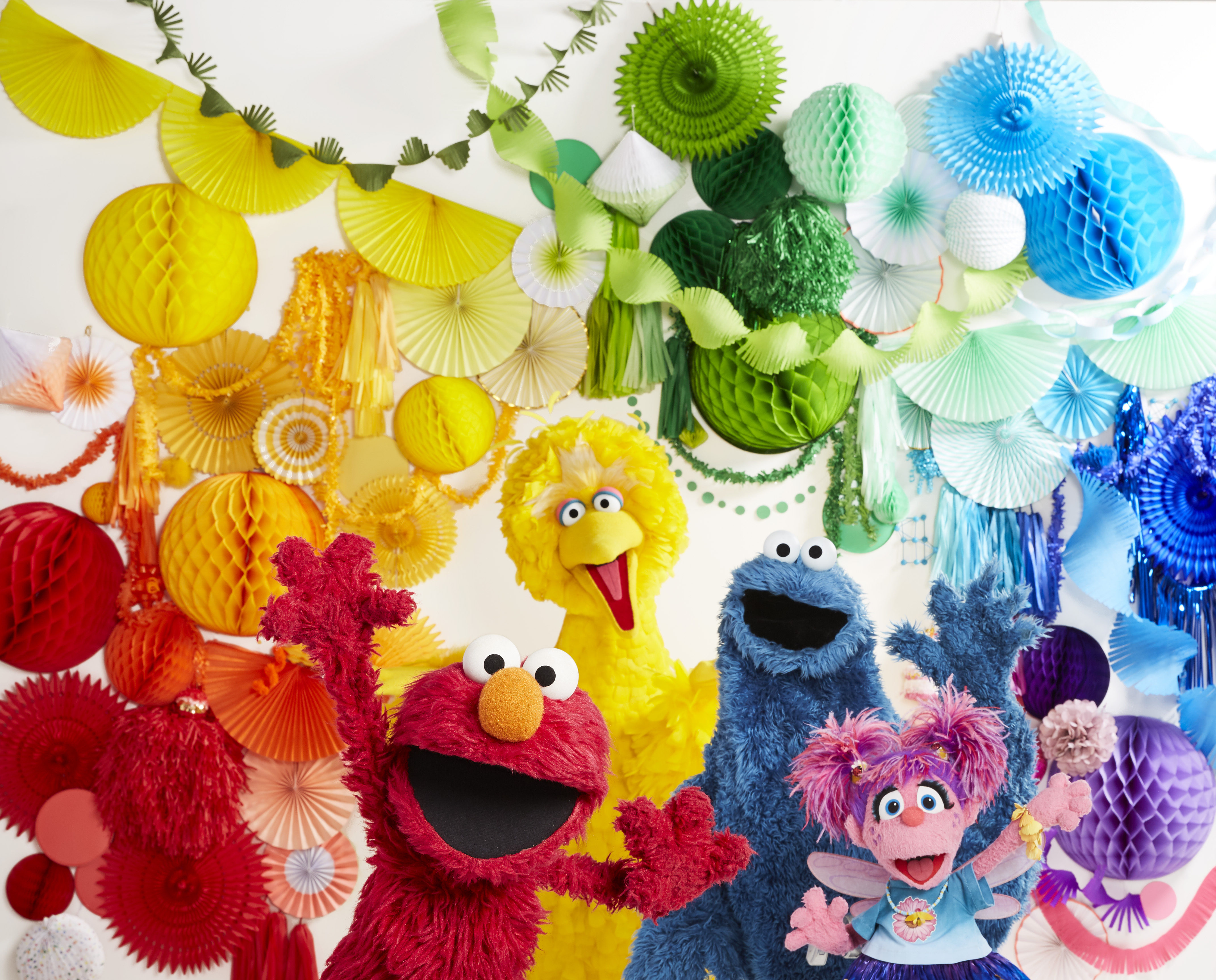 Elmo, Big Bird, Cookie Monster, Abby celebrate in front of paper decoration in rainbow colors.