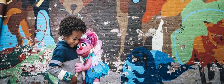 Abby hugs a child in front of a brick wall with a colorful, abstract mural.