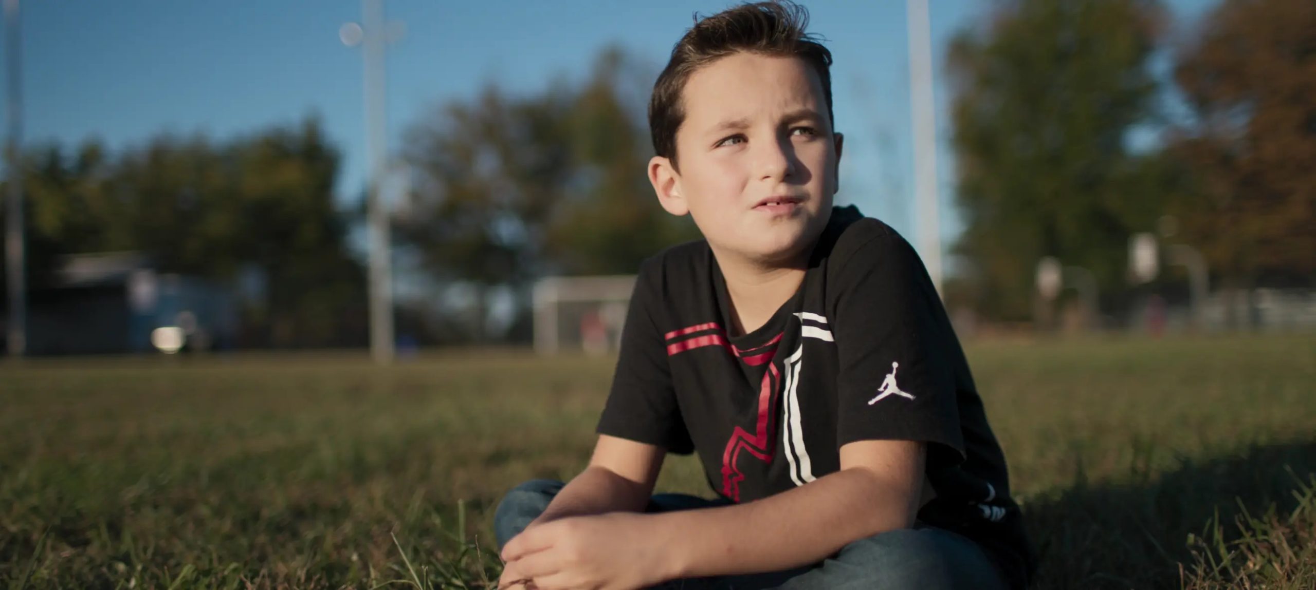 A young boy sits on the grass in a soccer field. He looks off into the distance.