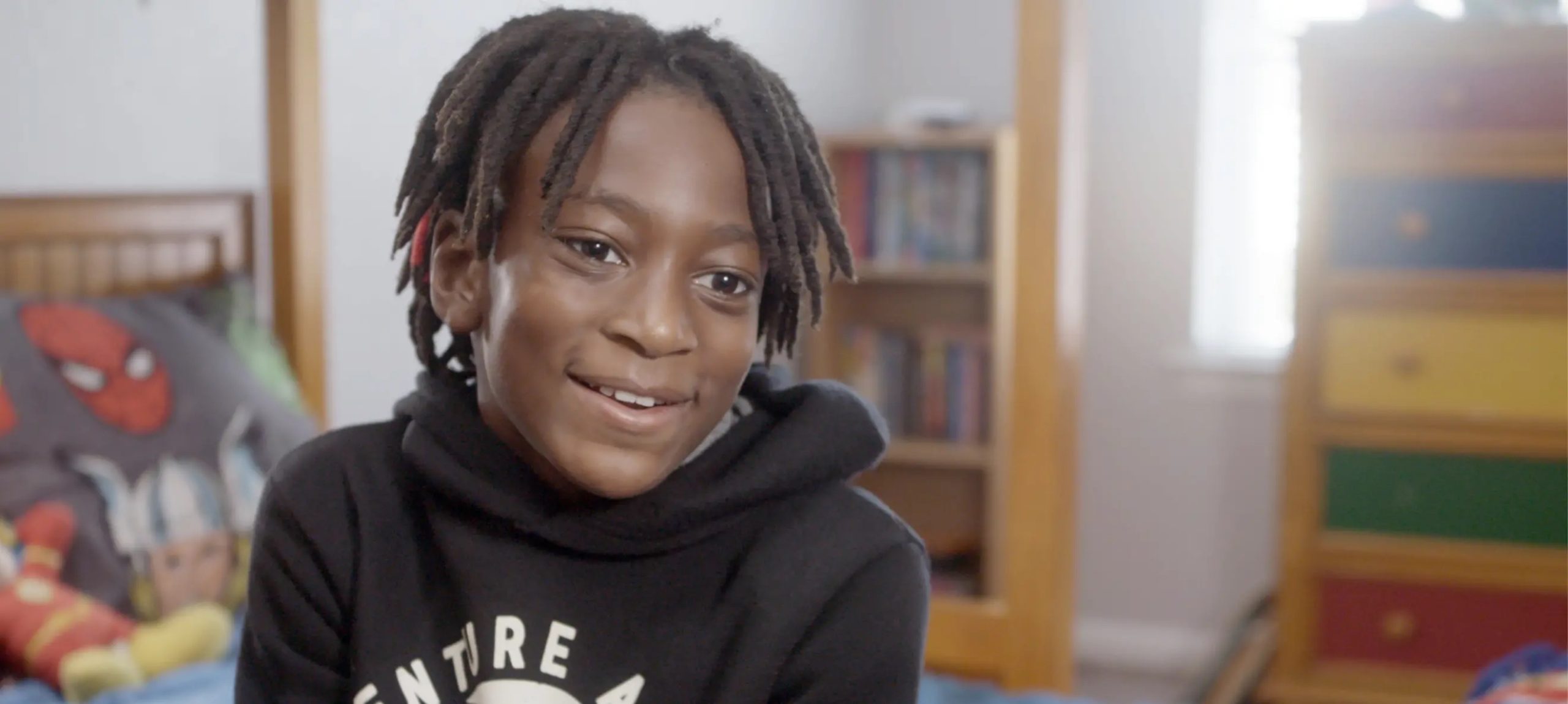 A young boy sits in a playroom and smiles at the camera.