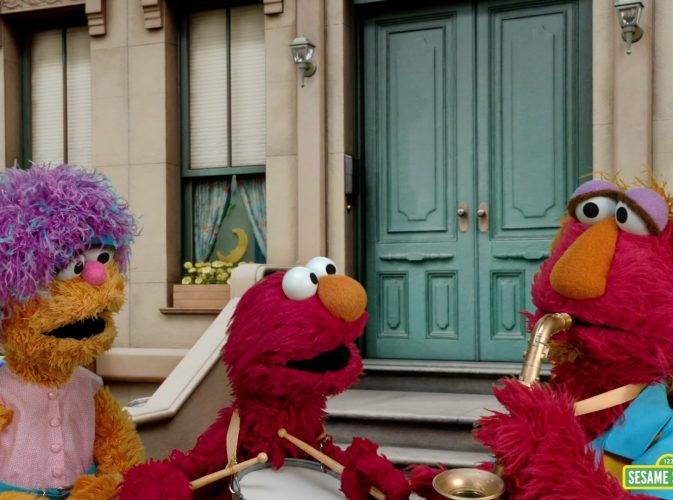Elmo playing a drum, standing with his mom and Dad. His father is playing a saxophone.