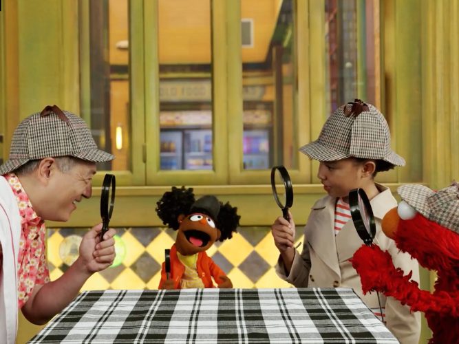 A group of detectives sit around a table with Gabrielle and Elmo